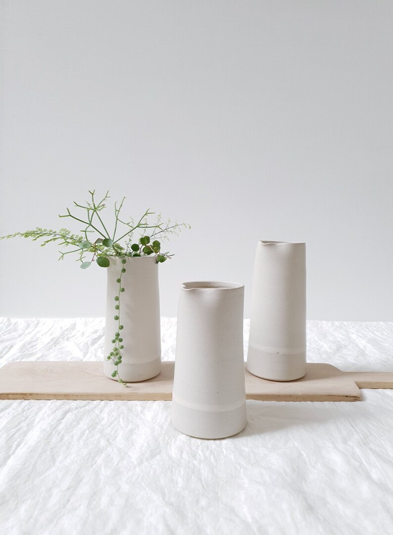 May include: Three white ceramic vases, two with a slightly flared lip and one with a straight lip, on a wooden surface. The vase in the center has a sprig of green foliage.
