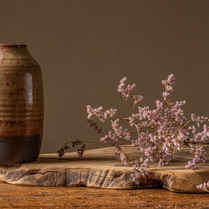 May include: A rustic brown ceramic vase with a dark brown rim sits on a wooden slab with dried pink flowers.