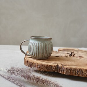 Puede incluir: Una taza de cerámica con detalles acanalados verticales y un esmalte azul claro. La taza tiene una forma redondeada con un asa pequeña. La taza se exhibe sobre una losa de madera con borde natural y unas ramitas de hierba ornamental seca.