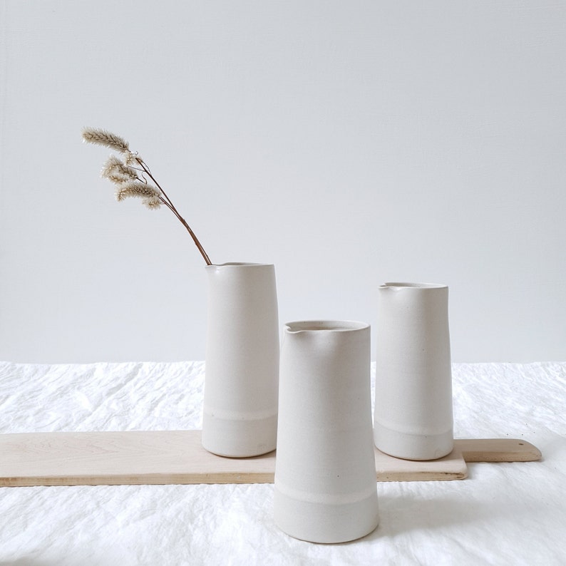 May include: Three white ceramic pitchers on a wooden board. The pitchers are all the same shape, but different sizes. The largest pitcher is in the background, and the smallest pitcher is in the foreground. The pitchers are all on a white linen tablecloth.