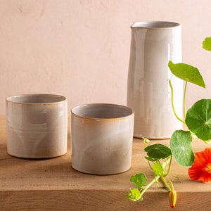 May include: Three ceramic cups and a pitcher, all in a light gray color, sit on a wooden surface. A single orange flower and green leaves are in the foreground.