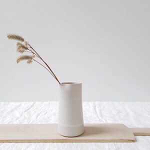 May include: A white ceramic pitcher with a slightly flared top and a white rim. The pitcher is sitting on a wooden cutting board and has a sprig of dried beige flowers in it.