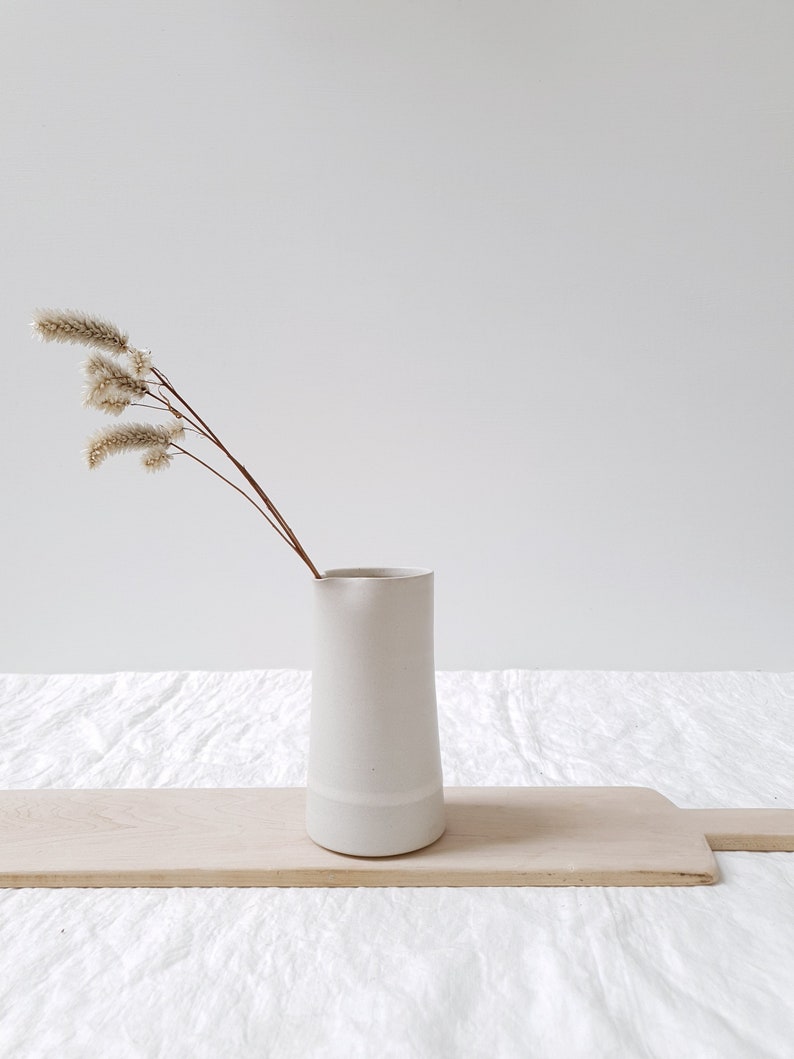 May include: A white ceramic pitcher with a slightly flared top and a white rim. The pitcher is sitting on a wooden cutting board and has a sprig of dried beige flowers in it.