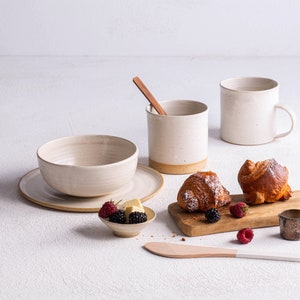 May include: A white ceramic bowl with a white ceramic plate, two white ceramic mugs, a wooden cutting board with two croissants, a small white bowl with raspberries and blackberries, a wooden spoon, and a small metal cup.