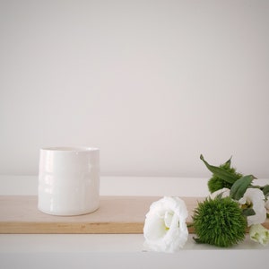 Op de afbeelding: Een witte keramische beker op een houten plank met witte bloemen en groen blad.