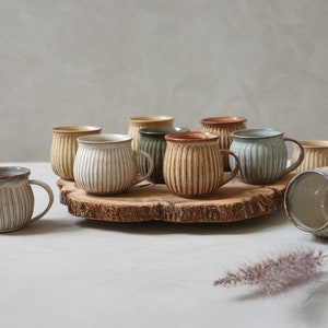 Może przedstawiać: Zbiór dziesięciu ceramicznych kubków z żebrowanym wzorem w różnych odcieniach brązu, beżu i błękitu. Kubki są ułożone na drewnianej powierzchni.