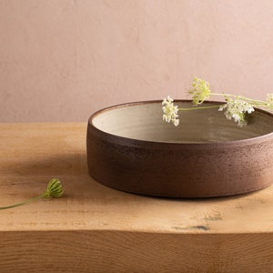 Op de afbeelding: Een bruine keramische kom met een witte binnenkant staat op een houten oppervlak. Witte bloemen en groene bladeren zijn rond de kom gerangschikt.