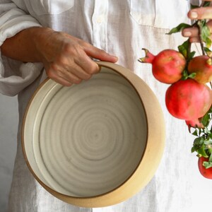 May include: A large, round, white ceramic bowl with a brown rim. The bowl has a spiral pattern on the inside. A hand is holding the bowl. A branch of red pomegranates with green leaves is hanging down.