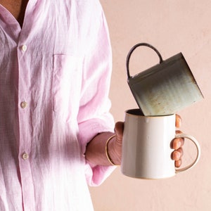 May include: A person pouring liquid from a light green ceramic mug into a white ceramic mug with a gold rim.