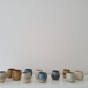 May include: A collection of 14 small, hand-thrown ceramic cups in various shades of brown, blue, and white. The cups are arranged in a row on a white surface.