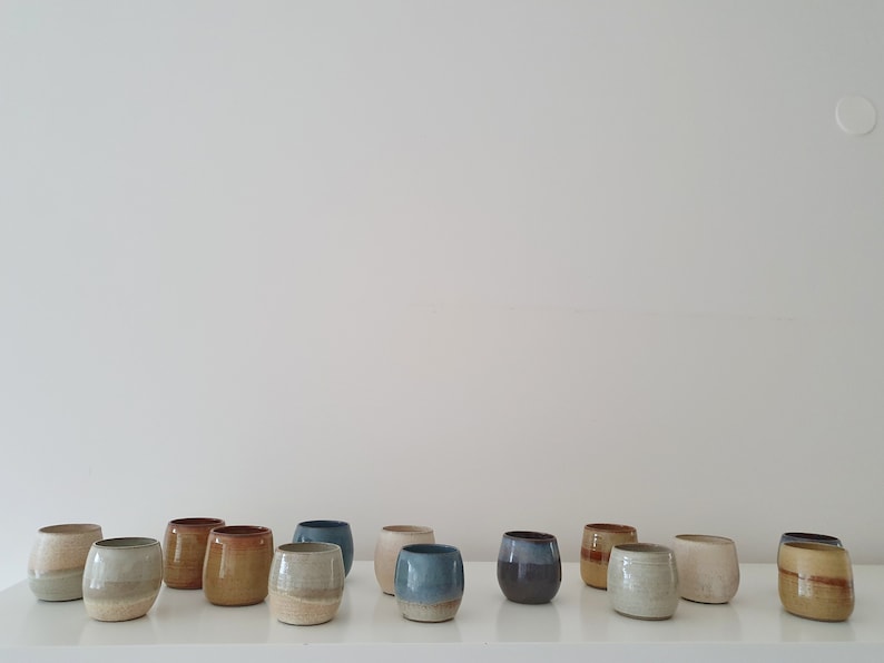 May include: A collection of 14 small, hand-thrown ceramic cups in various shades of brown, blue, and white. The cups are arranged in a row on a white surface.
