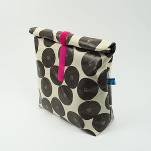 Könnte beinhalten: Eine Rolltop-Snacktasche mit cremefarbenem Hintergrund und einem Muster aus dunkelgrauen Kreisen, die kleine weiße Punkte enthalten. Die Tasche wird mit einem leuchtend pinken Klettverschlussband verschlossen und hat ein kleines blaues Etikett.
