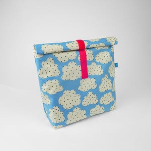 Op de afbeelding: Een blauwe lunchtas met een wolkenpatroon. De wolken zijn wit met zwarte stippen. Een felroze band sluit de opgerolde bovenkant. De tas heeft een klein blauw label aan de zijkant.