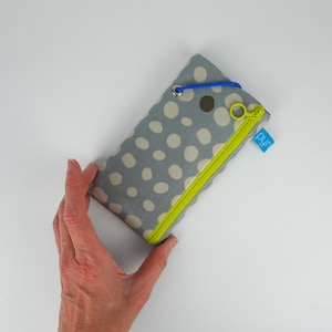 Può includere: Un astuccio in tessuto grigio con pois bianchi, una cerniera verde brillante e un anello elastico blu. L'astuccio ha una piccola etichetta blu con la scritta "pyn".