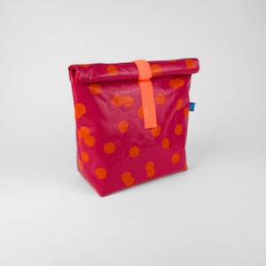 Op de afbeelding: Een magenta lunchtas met oranje stippen. De tas heeft een oprolbare sluiting die wordt vastgezet met een oranje band. Een klein blauw label is bevestigd. Ontworpen voor het vervoeren van voedsel.