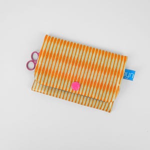 Könnte beinhalten: Eine rechteckige Stofftasche mit einem sich wiederholenden orangefarbenen und beigen vertikalen Wellenmuster. Die Tasche wird mit einem leuchtend rosa Druckknopf verschlossen, enthält ein kleines blaues Etikett mit weißem Text und eine lila Schere.