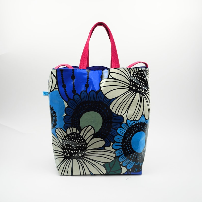 May include: A tote bag featuring a floral design in blue, white, and black. It has a bright pink handle and trim. The flowers are large and stylized, creating a striking pattern. The bag is suitable for carrying various items.