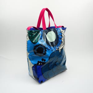 May include: A tote bag featuring a floral design in blue, white, and black. It has bright pink handles and a blue interior. The bag is made from a shiny, possibly waterproof material.