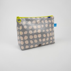 Könnte beinhalten: Graue Tasche mit einem Muster aus großen, hellbeigen Polka-Dots. Sie hat einen neongelben Reißverschluss und ein kleines blaues Etikett. Die Tasche ist wahrscheinlich für Kosmetika oder kleine Gegenstände gedacht.