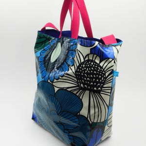 May include: A tote bag featuring a bold floral design in blue, white, and black. It has bright pink handles and a blue interior. The bag is constructed from a shiny, waterproof material.