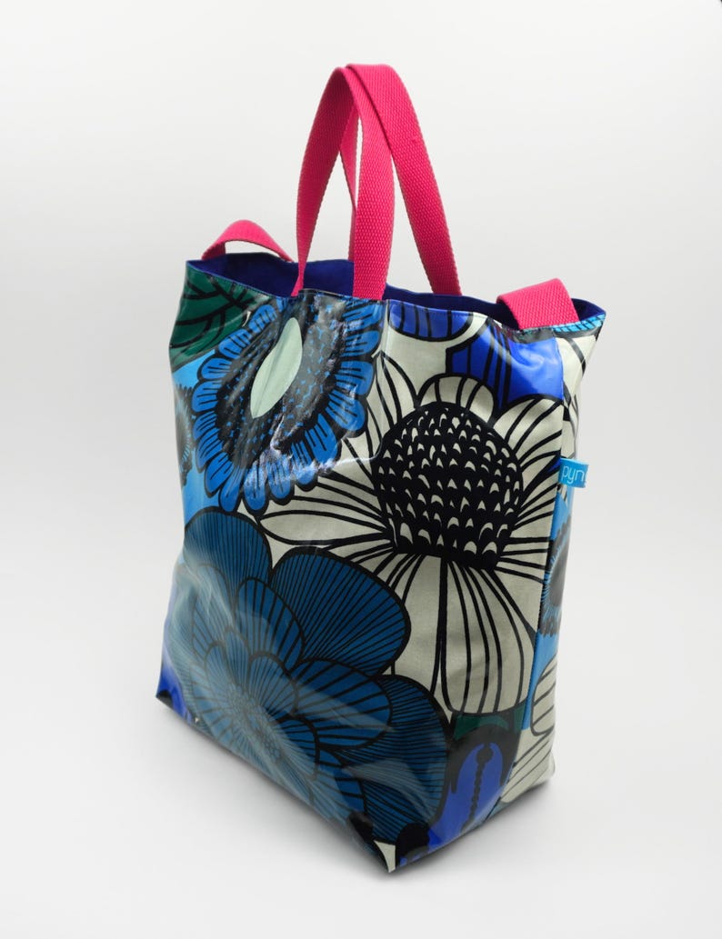 May include: A tote bag featuring a bold floral design in blue, white, and black. It has bright pink handles and a blue interior. The bag is constructed from a shiny, waterproof material.