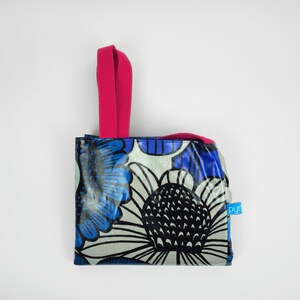 May include: A small tote bag featuring a bold floral design in blue, black, and white. It has a bright pink handle and a small blue tag with white text. The bag is designed for carrying items.