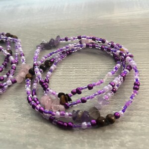 Puede incluir: Varias hebras de cuentas de cintura moradas, lavanda y transparentes con peque&ntilde;os detalles de piedras preciosas. Las cuentas est&aacute;n ensartadas, creando un efecto en capas. Las cuentas de cintura se muestran sobre una superficie gris claro.