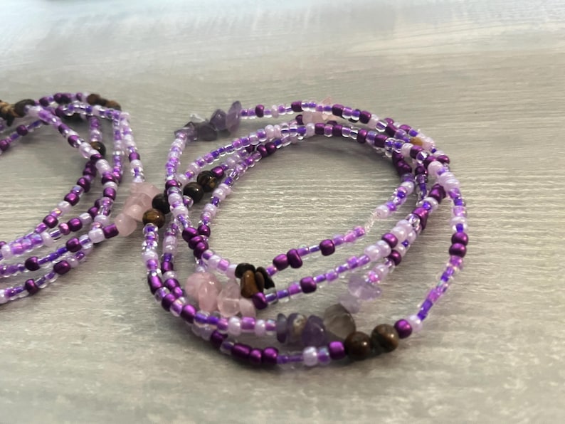 Puede incluir: Varias hebras de cuentas de cintura moradas, lavanda y transparentes con peque&ntilde;os detalles de piedras preciosas. Las cuentas est&aacute;n ensartadas, creando un efecto en capas. Las cuentas de cintura se muestran sobre una superficie gris claro.