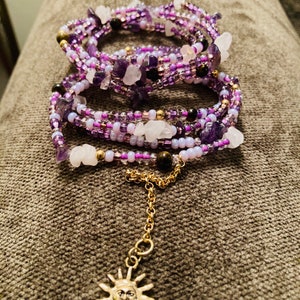 Puede incluir: Una pulsera envolvente con m&uacute;ltiples hilos de cuentas moradas y blancas, trozos de amatista y cuarzo, y detalles dorados. Una cadena dorada con un dije de sol cuelga de la pulsera. La pulsera se muestra sobre una superficie gris.