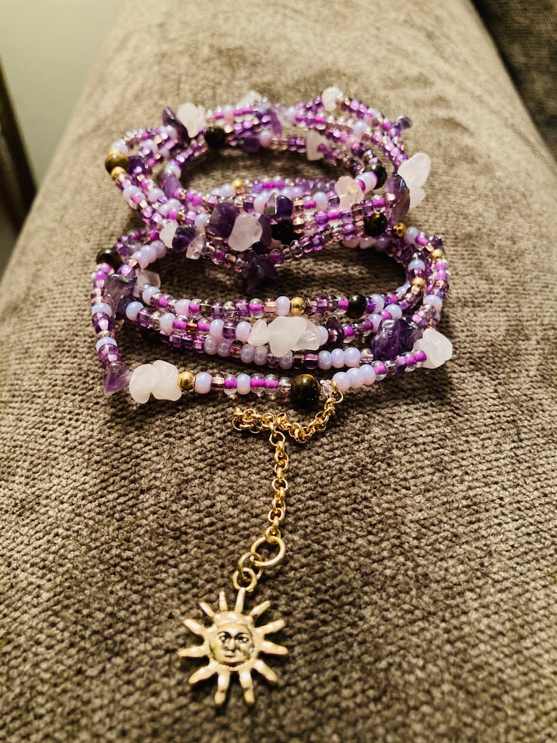 Puede incluir: Una pulsera envolvente con m&uacute;ltiples hilos de cuentas moradas y blancas, trozos de amatista y cuarzo, y detalles dorados. Una cadena dorada con un dije de sol cuelga de la pulsera. La pulsera se muestra sobre una superficie gris.