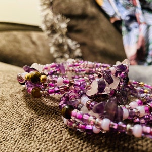 Puede incluir: Una colecci&oacute;n de pulseras de cuentas en tonos morados, rosas y blancos. Las pulseras presentan peque&ntilde;as cuentas, piezas de piedras preciosas m&aacute;s grandes y detalles dorados. Las joyas se exhiben sobre una superficie texturizada de tono neutro.