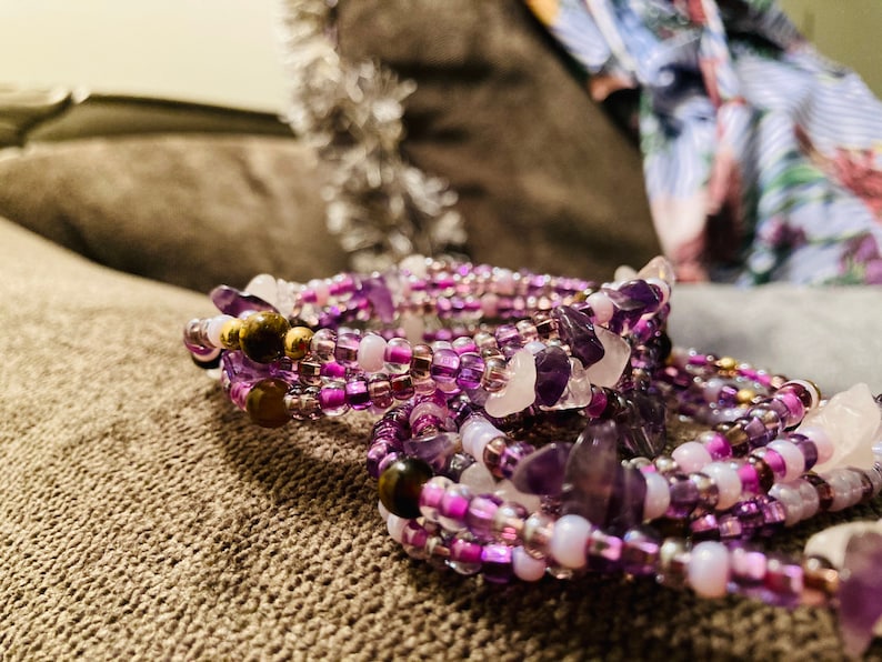 Puede incluir: Una colecci&oacute;n de pulseras de cuentas en tonos morados, rosas y blancos. Las pulseras presentan peque&ntilde;as cuentas, piezas de piedras preciosas m&aacute;s grandes y detalles dorados. Las joyas se exhiben sobre una superficie texturizada de tono neutro.