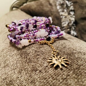 Puede incluir: Una pulsera de cuentas de varios hilos en tonos morado, blanco y dorado. La pulsera presenta una cadena dorada con un dije de sol. Las cuentas son una mezcla de peque&ntilde;as cuentas de semillas y piedras m&aacute;s grandes.