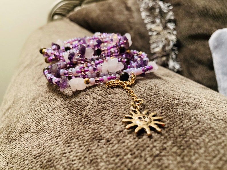 Puede incluir: Una pulsera de cuentas de varios hilos en tonos morado, blanco y dorado. La pulsera presenta una cadena dorada con un dije de sol. Las cuentas son una mezcla de peque&ntilde;as cuentas de semillas y piedras m&aacute;s grandes.