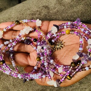 Puede incluir: Cintura de cuentas en tonos morados, lavanda y blanco, acentuada con cuentas doradas y un dije de sol. Las cuentas est&aacute;n intercaladas con peque&ntilde;as piedras preciosas y cristales, creando un efecto en capas.
