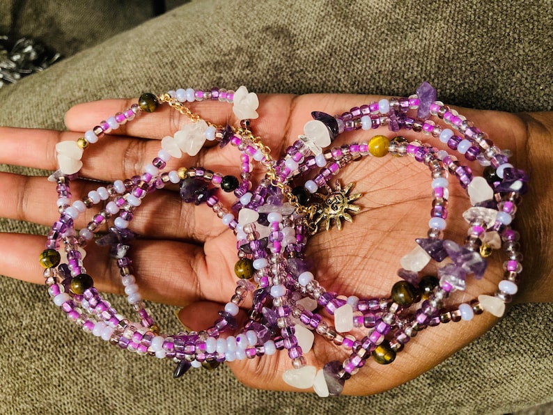 Puede incluir: Cintura de cuentas en tonos morados, lavanda y blanco, acentuada con cuentas doradas y un dije de sol. Las cuentas est&aacute;n intercaladas con peque&ntilde;as piedras preciosas y cristales, creando un efecto en capas.