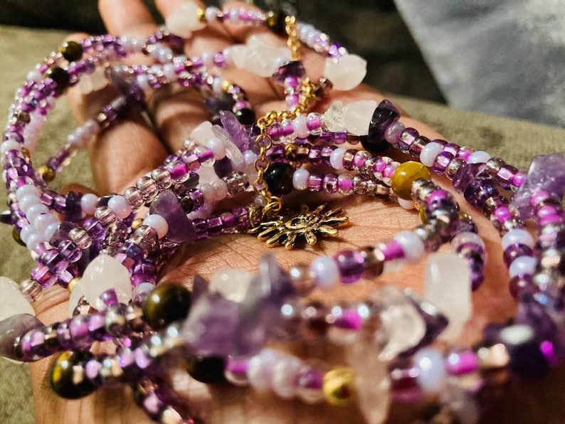 Puede incluir: Collar de cuentas de m&uacute;ltiples hilos con piedras de amatista, cuarzo transparente y ojo de tigre. El collar tiene detalles en morado, blanco y dorado, con un dije de sol dorado. Las cuentas son peque&ntilde;as y delicadas.