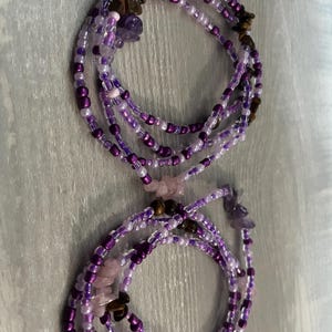 Puede incluir: Dos hilos de cuentas de cintura moradas con una mezcla de peque&ntilde;as cuentas y acentos de piedras preciosas m&aacute;s grandes. Las cuentas son en tonos de morado, lavanda y blanco. Las piedras preciosas incluyen amatista y ojo de tigre. Los hilos est&aacute;n dispuestos en un patr&oacute;n circular.