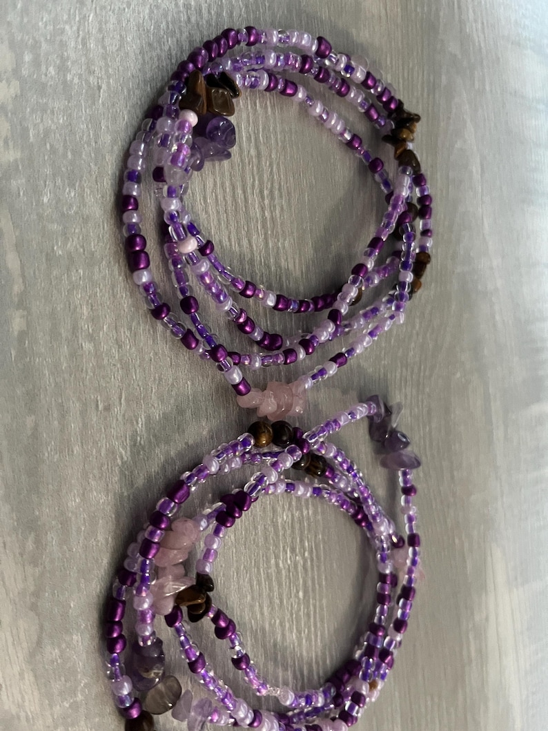 Puede incluir: Dos hilos de cuentas de cintura moradas con una mezcla de peque&ntilde;as cuentas y acentos de piedras preciosas m&aacute;s grandes. Las cuentas son en tonos de morado, lavanda y blanco. Las piedras preciosas incluyen amatista y ojo de tigre. Los hilos est&aacute;n dispuestos en un patr&oacute;n circular.