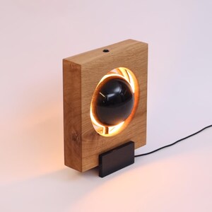 Könnte beinhalten: Eine moderne Holzlampe mit einem rechteckigen Eichenrahmen. Die Lampe hat eine schwarze Kugel in der Mitte, die von einem warmen, orangefarbenen Licht beleuchtet wird. Ein kleiner schwarzer Sockel und ein schwarzes Netzkabel sind angebracht.