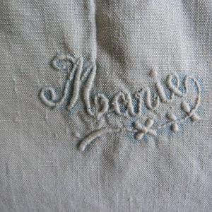 May include: Close-up of a light blue fabric with the embroidered name "Marie" in a cursive script. The embroidery is in a lighter shade of blue and includes small floral accents.