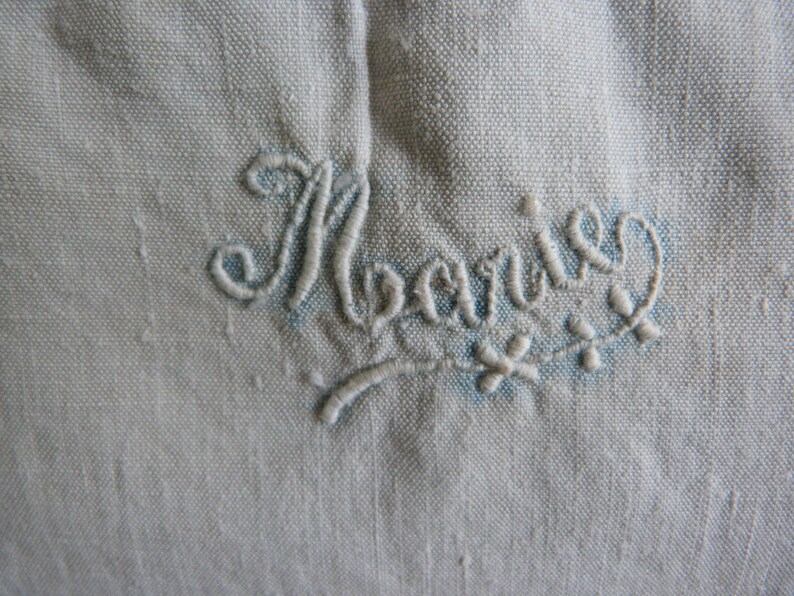 May include: Close-up of a light blue fabric with the embroidered name "Marie" in a cursive script. The embroidery is in a lighter shade of blue and includes small floral accents.