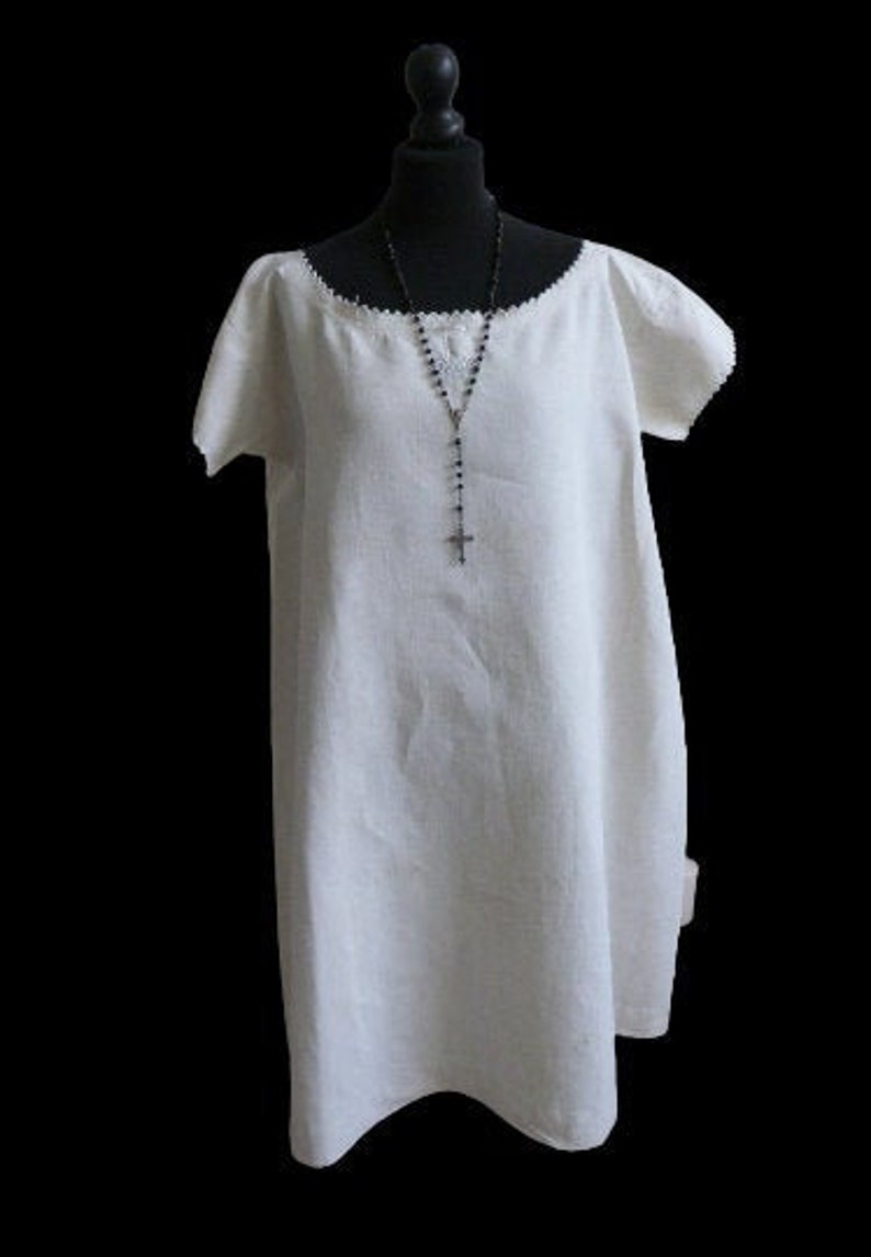 May include: A white linen dress with short sleeves and a scoop neckline, displayed on a black mannequin. A black rosary with a cross hangs from the neckline. The dress has a simple, flowing design.