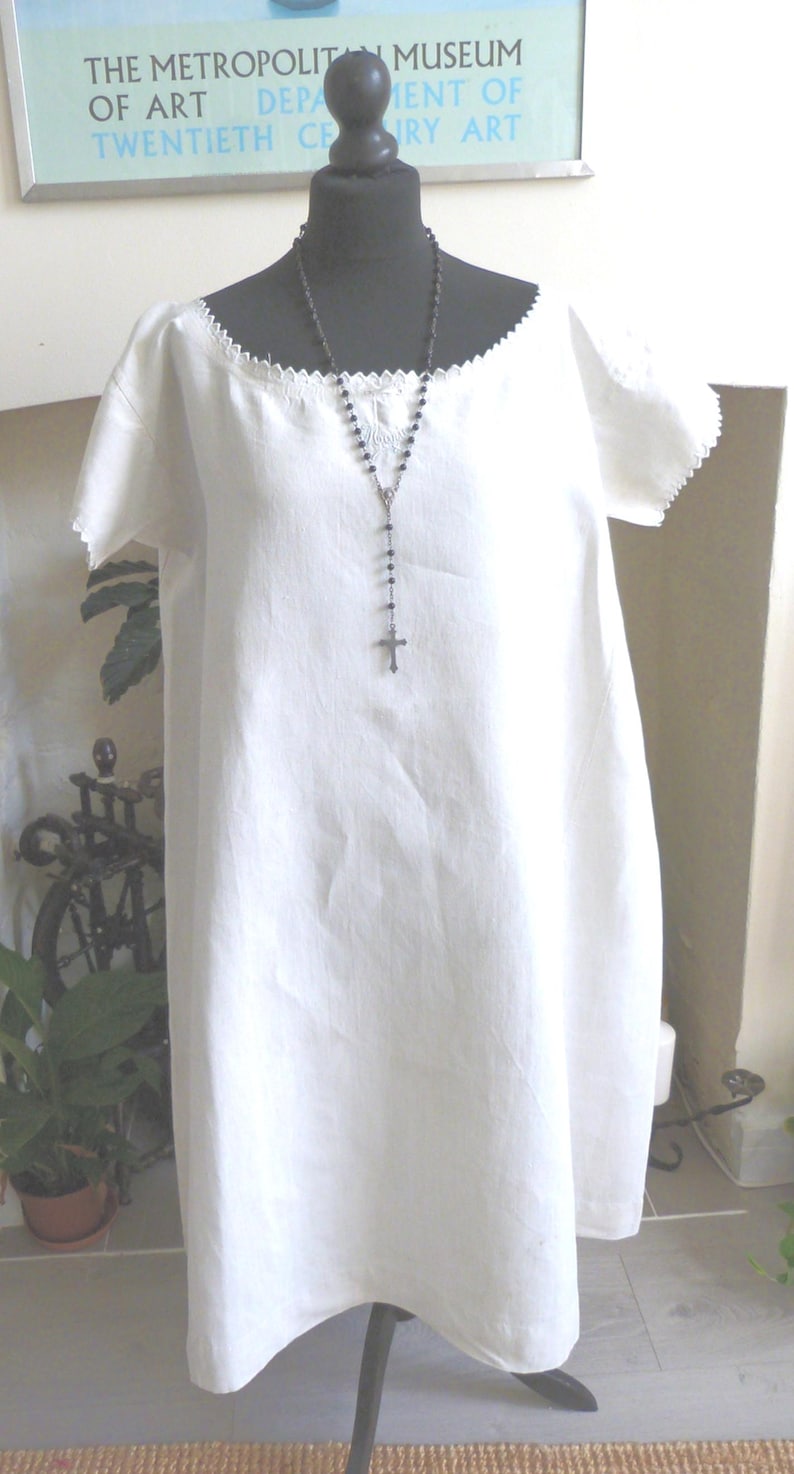 May include: A white linen dress with short sleeves and a decorative neckline is displayed on a black mannequin. A silver rosary hangs around the mannequin's neck. The dress is knee-length and appears to be handmade.