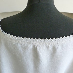 May include: White linen top with a scalloped neckline. The neckline is trimmed with a delicate, repeating pattern. The top is displayed on a black mannequin, showcasing the garment's shape and details. The fabric appears soft and slightly textured.
