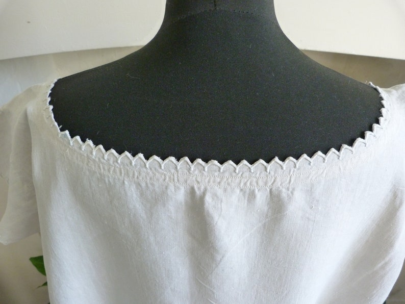 May include: White linen top with a scalloped neckline. The neckline is trimmed with a delicate, repeating pattern. The top is displayed on a black mannequin, showcasing the garment's shape and details. The fabric appears soft and slightly textured.