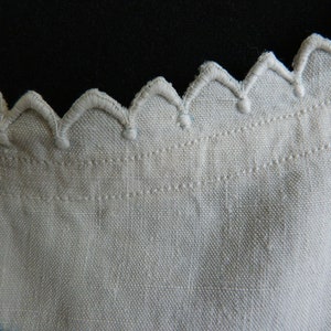 May include: Close-up of a cream-colored fabric with a scalloped edge. The edge is embroidered with a repeating pattern of rounded peaks. The fabric has a textured appearance, with visible woven threads and a subtle sheen.