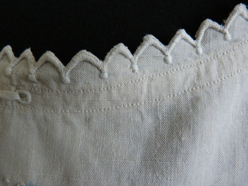 May include: Close-up of a cream-colored fabric with a scalloped edge. The edge is embroidered with a repeating pattern of rounded peaks. The fabric has a textured appearance, with visible woven threads and a subtle sheen.