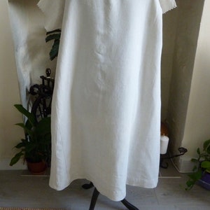 May include: A white linen dress with a scalloped neckline and short sleeves. The dress is displayed on a black metal stand. The dress appears to be knee-length and has a simple, flowing design.