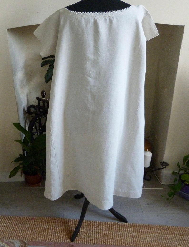 May include: A white linen dress with a scalloped neckline and short sleeves. The dress is displayed on a black metal stand. The dress appears to be knee-length and has a simple, flowing design.